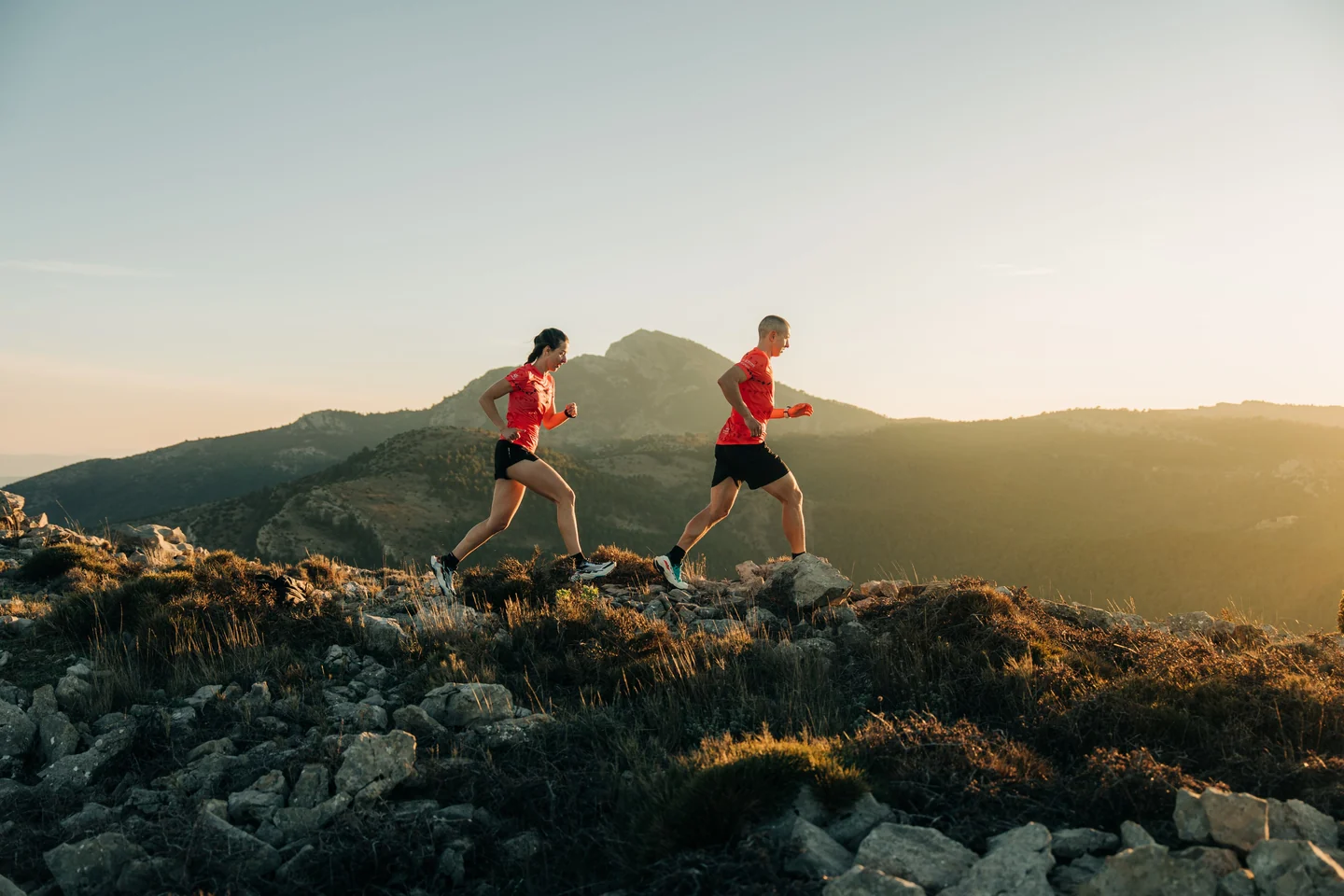 Dos corredores con camisetas rojas avanzando por una cresta de montaña al atardecer